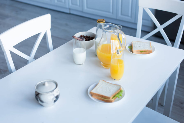 sándwiches, copos de maíz crujientes, leche y jugo de naranja servidos en la mesa de la cocina - Foto, imagen