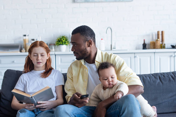 african american man holding tv remote controller near daughter sitting with book with closed eyes - 写真・画像