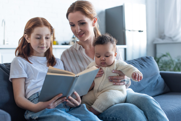 sorridente ragazza lettura libro vicino madre e neonato africano americano sorella - Foto, immagini