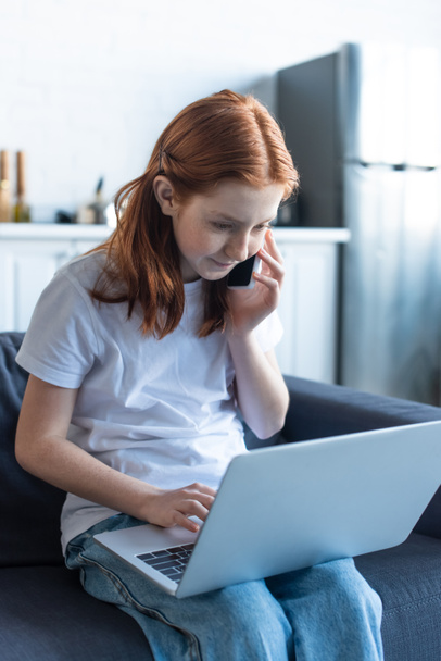 preteen girl with red hair using laptop and talking on smartphone at home - Foto, Imagem