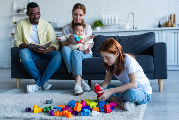 multiethnic couple sitting on couch with infant near preteen daughter playing with building blocks - Photo, Image