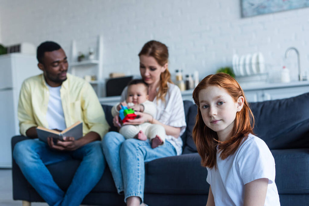 redhead preteen girl looking at camera near multiethnic family sitting on sofa on blurred background - Photo, Image