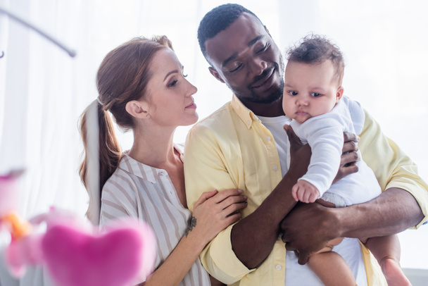 pleased interracial parents looking at baby on blurred foreground - Foto, immagini