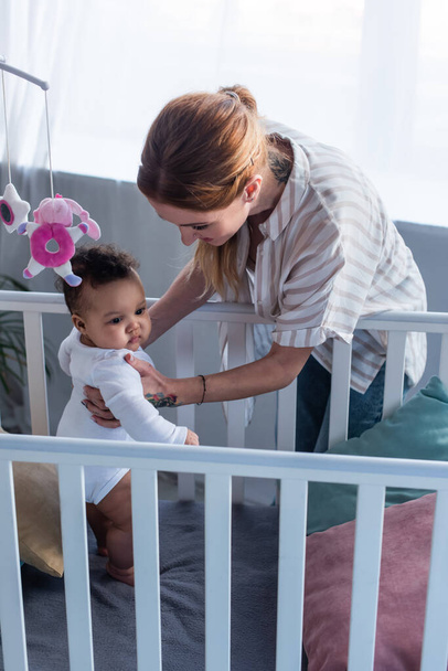 mother supporting little african american daughter standing in crib - Photo, Image