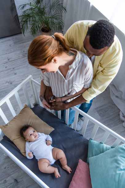 overhead view of african american man embracing wife near infant kid lying in crib - Фото, зображення