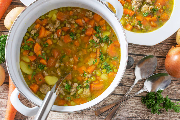 Vegetable soup with minced meat made with green peas, carrots, potatoes and topped with parsley. Served in a pot with ladle on rustic wooden table background. Overhead view - Photo, Image