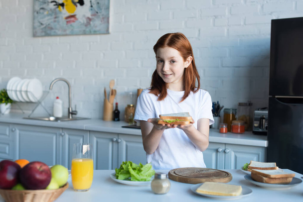 redhead preteen girl holding tasty sandwich in kitchen - Fotó, kép