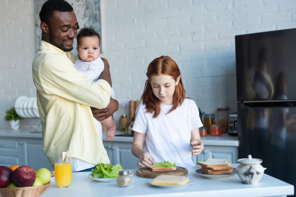 щасливий афроамериканський чоловік з немовлям дивиться на дочку готує бутерброди
 - Фото, зображення
