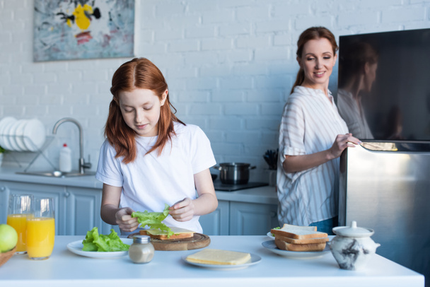pleased woman looking at daughter preparing sandwiches for breakfast - Fotó, kép