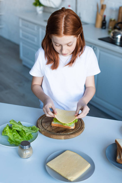 high angle view of redhead girl preparing sandwich with lettuce and cheese - Фото, изображение