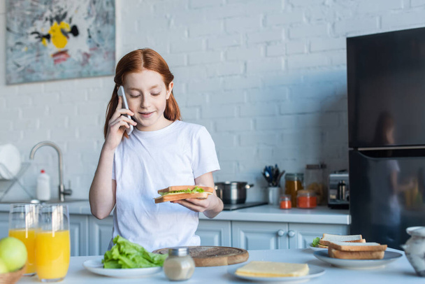 redhead girl holding sandwich and talking on cellphone in kitchen - Photo, Image