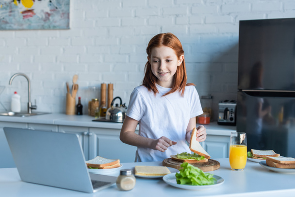 redhead girl looking at laptop while making sandwich for breakfast - Foto, Imagen