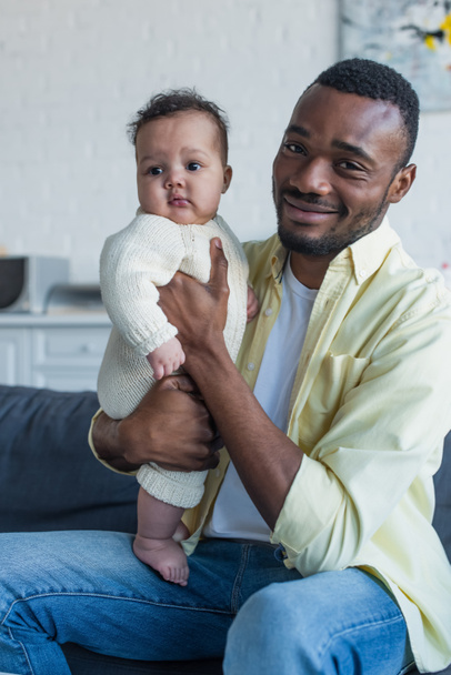 onnellinen afrikkalainen amerikkalainen mies katselee kameraa istuessaan sohvalla vauvan kanssa - Valokuva, kuva