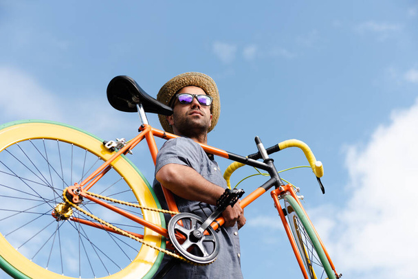 Latynoski hipster z kolorowym rowerem na ramieniu z niebieskim niebem jako tłem. Koncepcja zrównoważonej mobilności i transportu. - Zdjęcie, obraz