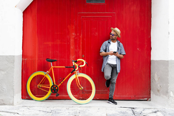 Латиноамериканец против старой красной двери, держа телефон рядом со своим винтажным велосипедом. Концепция устойчивого туризма и мобильности - Фото, изображение