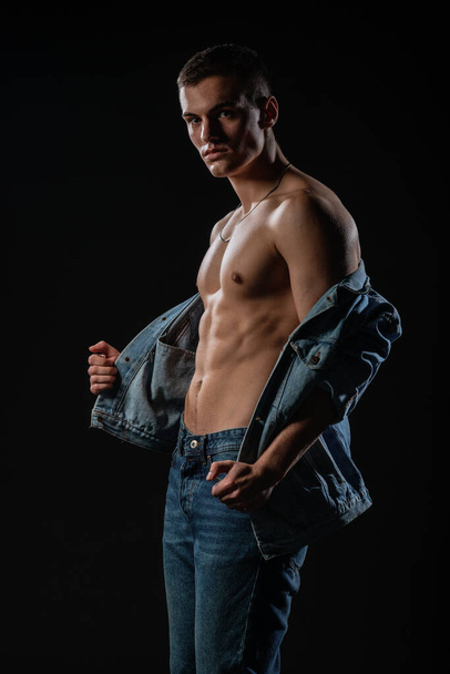Sexy man with a naked torso posing in blue jeans with a denim jacket at his waist - Photo, image