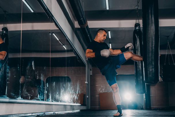 musculoso guapo luchador dando una fuerte patada hacia adelante durante una ronda de práctica con una bolsa de boxeo
 - Foto, Imagen