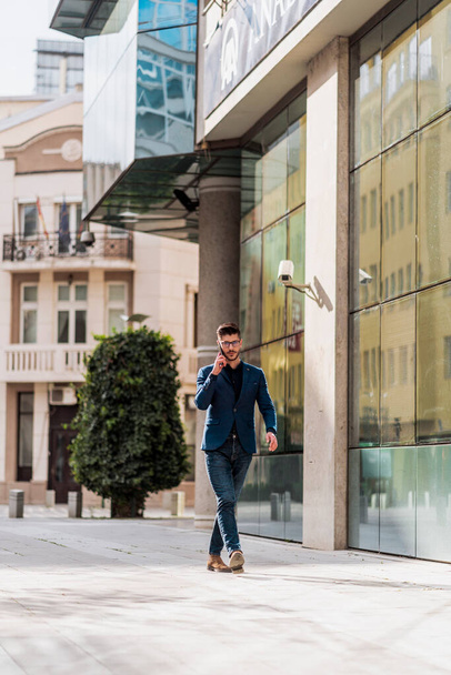 Atractivo hombre de negocios guapo de pie frente al edificio y hablando por teléfono móvil - Foto, Imagen