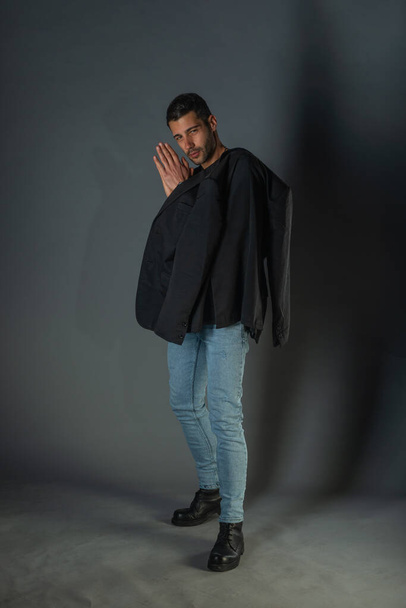 Full length trendy νεαρός άνδρας φορώντας μαύρο σακάκι και τζιν, στέκεται πάνω σε ένα γκρι φόντο - Φωτογραφία, εικόνα