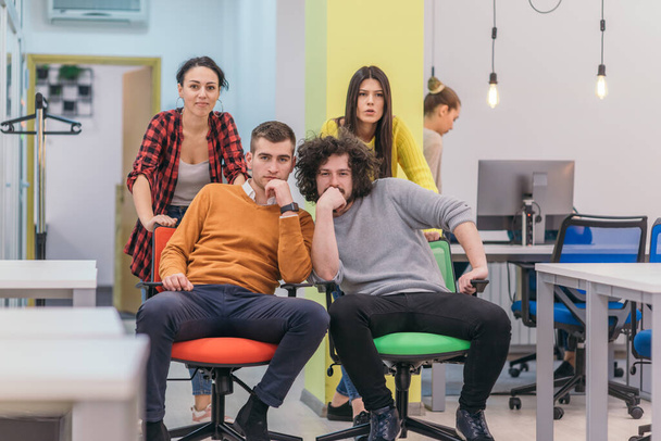 Cuatro jóvenes empresarios alegres en ropa casual inteligente que se divierten mientras compiten en sillas de oficina y sonriendo. - Foto, Imagen