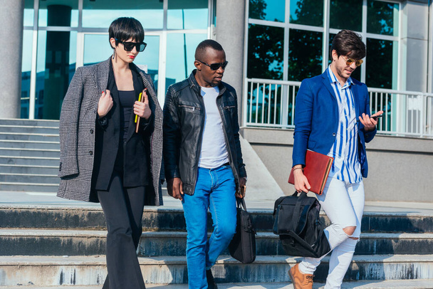 Jóvenes multiétnicos modernos en ropa formal discutiendo negocios mientras caminan fuera del edificio de oficinas - Foto, Imagen