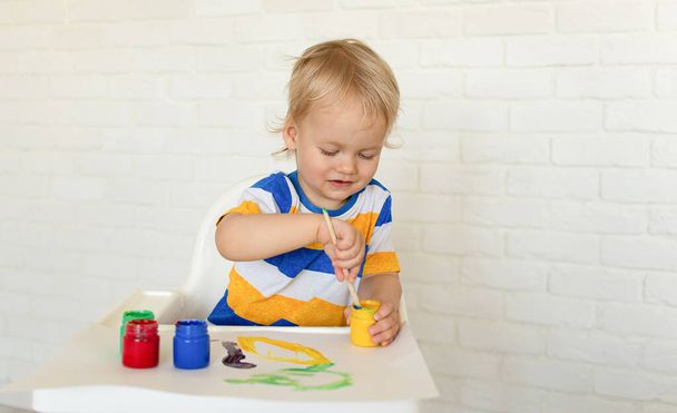 Kid draw with paints, playing at home sits at a kids table. Todler with educational toys for creativity. Boy play at home. Bright paint gouache stained face and hands of a child. - Photo, Image