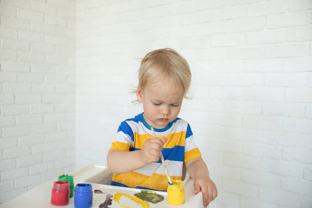 Kid draw with paints, playing at home sits at a kids table. Todler with educational toys for creativity. Boy play at home. Bright paint gouache stained face and hands of a child. - Foto, Imagen