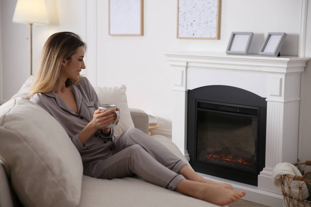 Young woman with cup of hot drink resting on sofa near fireplace at home - Foto, Imagem