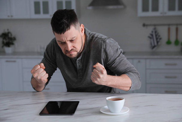 Emotional man with tablet at table in kitchen. Online hate concept - Photo, Image