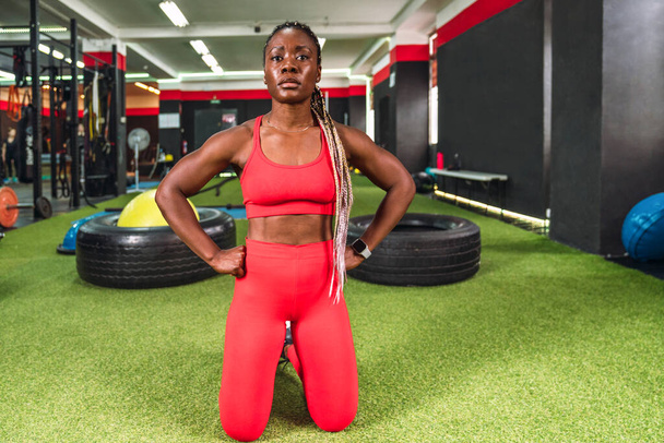 体育館にいる強いアスレチックブラックの女性は、準備ができてスポーツをするために膝に集中し、赤いスポーツウェアを着て - 写真・画像