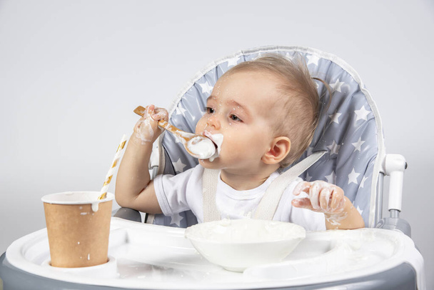 Little beautiful child eating a wooden spoon of yogurt from a plate and a paper eco cup with a straw with milk on a high gray chair on a white background - Photo, Image