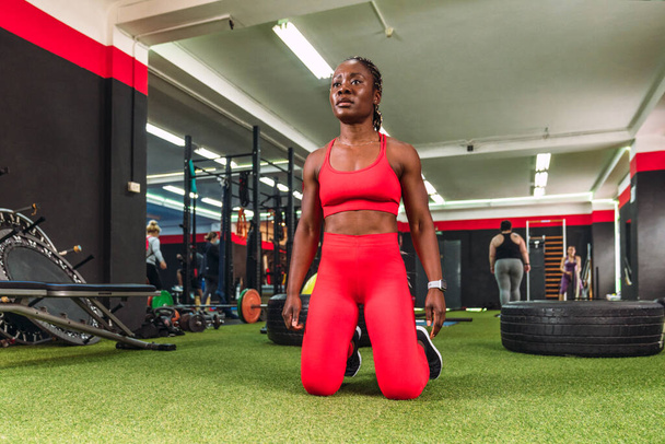 ισχυρή αθλητική μαύρη γυναίκα σε ένα γυμναστήριο, έτοιμη και συγκεντρωμένη στα γόνατά της για να κάνει σπορ, ντυμένη με κόκκινα αθλητικά ρούχα - Φωτογραφία, εικόνα