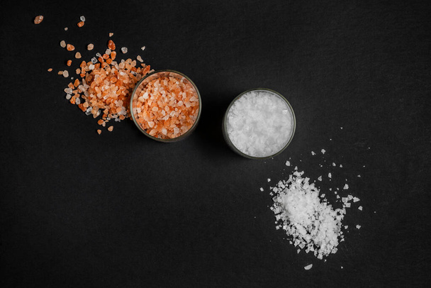 two glass containers from top view filled one with sea salt flakes and the other with himalayan salt rocks on black background - Photo, Image
