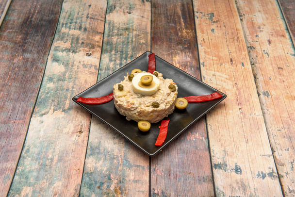 Типичная испанская тапа русского салата с вареным яйцом, оливками, красным перцем и вареной картошкой с майонезом на квадратной черной тарелке - Фото, изображение