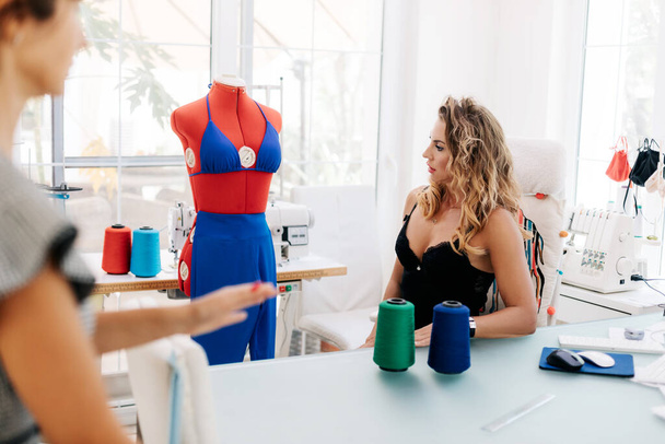 若いデザイナーが縫製工場のテーブルに座り、同僚と新しいファッションデザインについて話し合う - 写真・画像