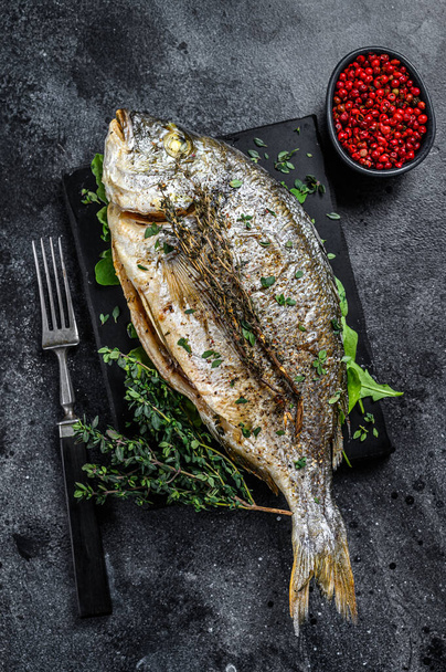 Roasted sea bream fish with herbs on a cutting board. Black background. Top view - Foto, Bild