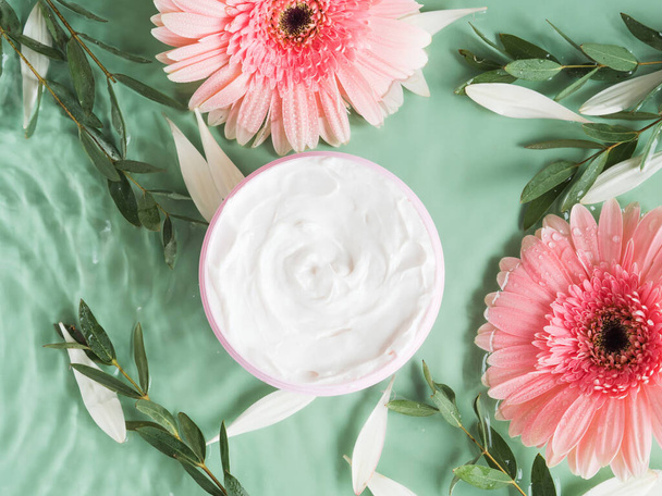 Skin care flat lay with generic cream jar with pink gerbera in water on green background - Foto, afbeelding