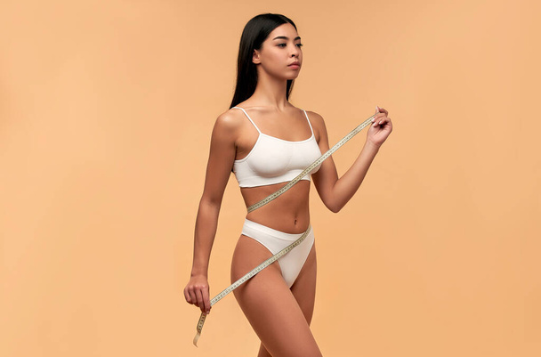 Young Asian woman in white lingerie with a toned body and a slender figure with a measuring tape on a beige background. Sports, healthy lifestyle. Spa body care. Plastic surgery. - Zdjęcie, obraz