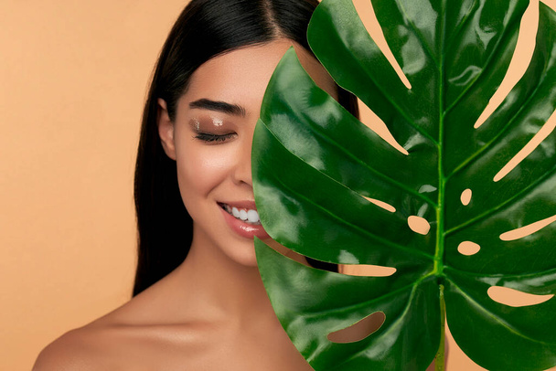 Young Asian woman with clean radiant face skin with green leaf on beige background. Spa care, facial skin care, beauty cosmetology. Natural cosmetics concept. - Valokuva, kuva