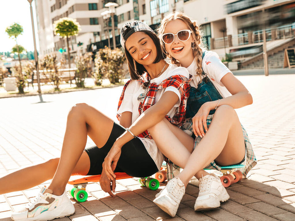 Two young smiling beautiful female with colorful penny skateboards. Women in summer hipster clothes sitting in the street background. Positive models having fun and going crazy - Zdjęcie, obraz