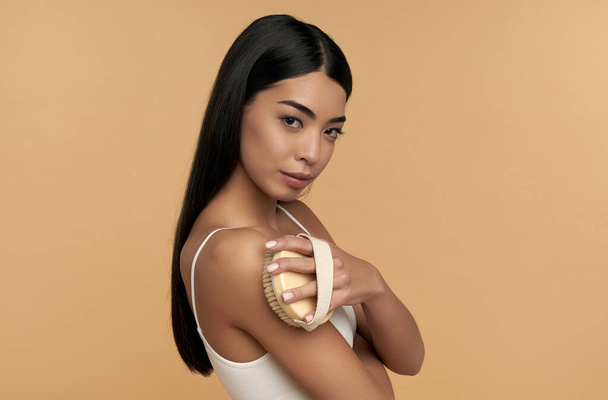 Jonge Aziatische vrouw in witte lingerie en schone stralende huid maakt anti-cellulitis massage met een droge borstel op een beige achtergrond. Lichaamsscrubbing, gewichtsverlies, lichaamsverzorging, spa-behandelingen. - Foto, afbeelding