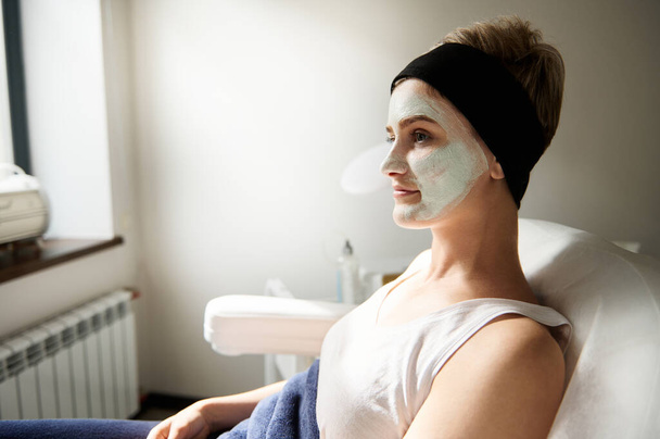Side portrait of a young woman sitting on a chair in spa salon with beauty cleansing mask on her face - Photo, Image
