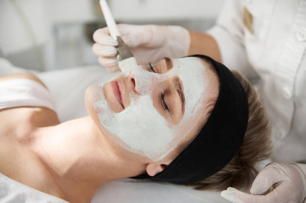 Крупный план косметолога, надевающего альгиновую маску на красивое женское лицо в курортной клинике - Фото, изображение