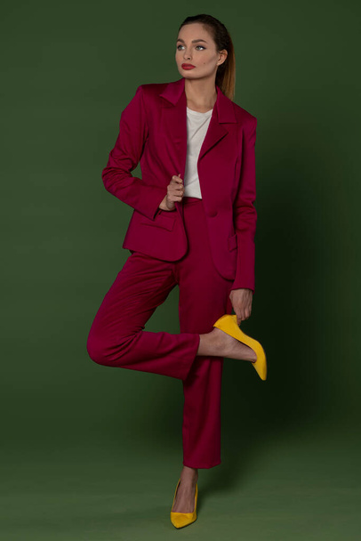 fashion model posing on green backgound , she dress a fuchsia suit with pants and jacket , and yellow high heel shoes - Photo, Image
