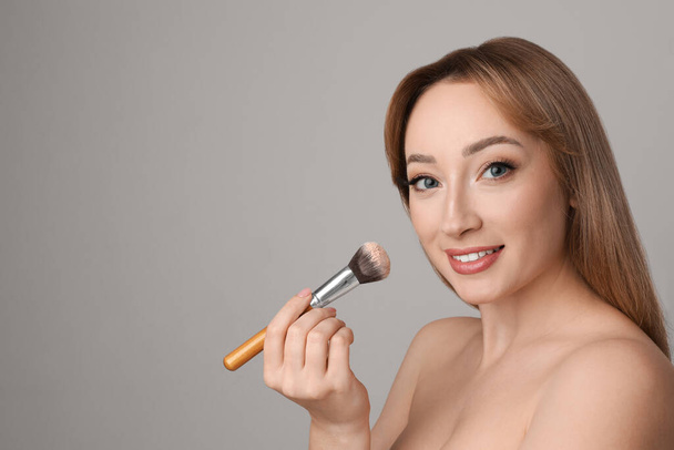 Beautiful young woman applying face powder with brush on grey background. Space for text - Fotografie, Obrázek