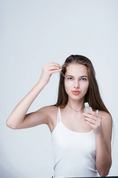 A charming girl in a white T-shirt with long hair holds a serum in her hands and applies it with a pipette to her face - Foto, afbeelding