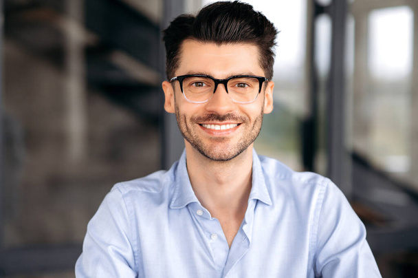 Close-up portret van knappe zelfverzekerde jonge blanke man met een bril, stijlvol shirt aan, op kantoor, direct naar de camera kijkend en lachend vriendelijk - Foto, afbeelding