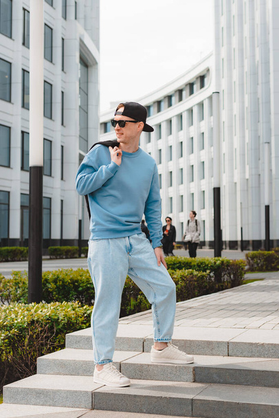 Young free beautiful fashionable hipster guy walking down the street, wearing a cap sunglasses, urban style, t-shirt, sport casual background modern white building - Zdjęcie, obraz