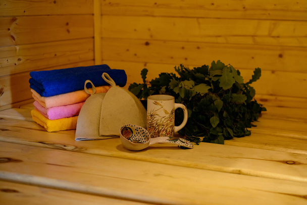 аксесуари для лазні: мітла, соломинка, банні капелюхи, різнокольорові рушники на дерев'яній полиці
 - Фото, зображення