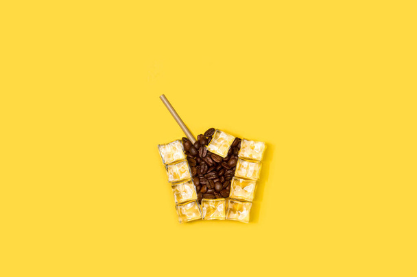 Ice cubes and coffee beans drinking glass shaped on a yellow background - Fotó, kép